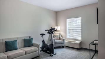A room with a sofa, an exercise bike, a chair, a table, and a window with blinds.
