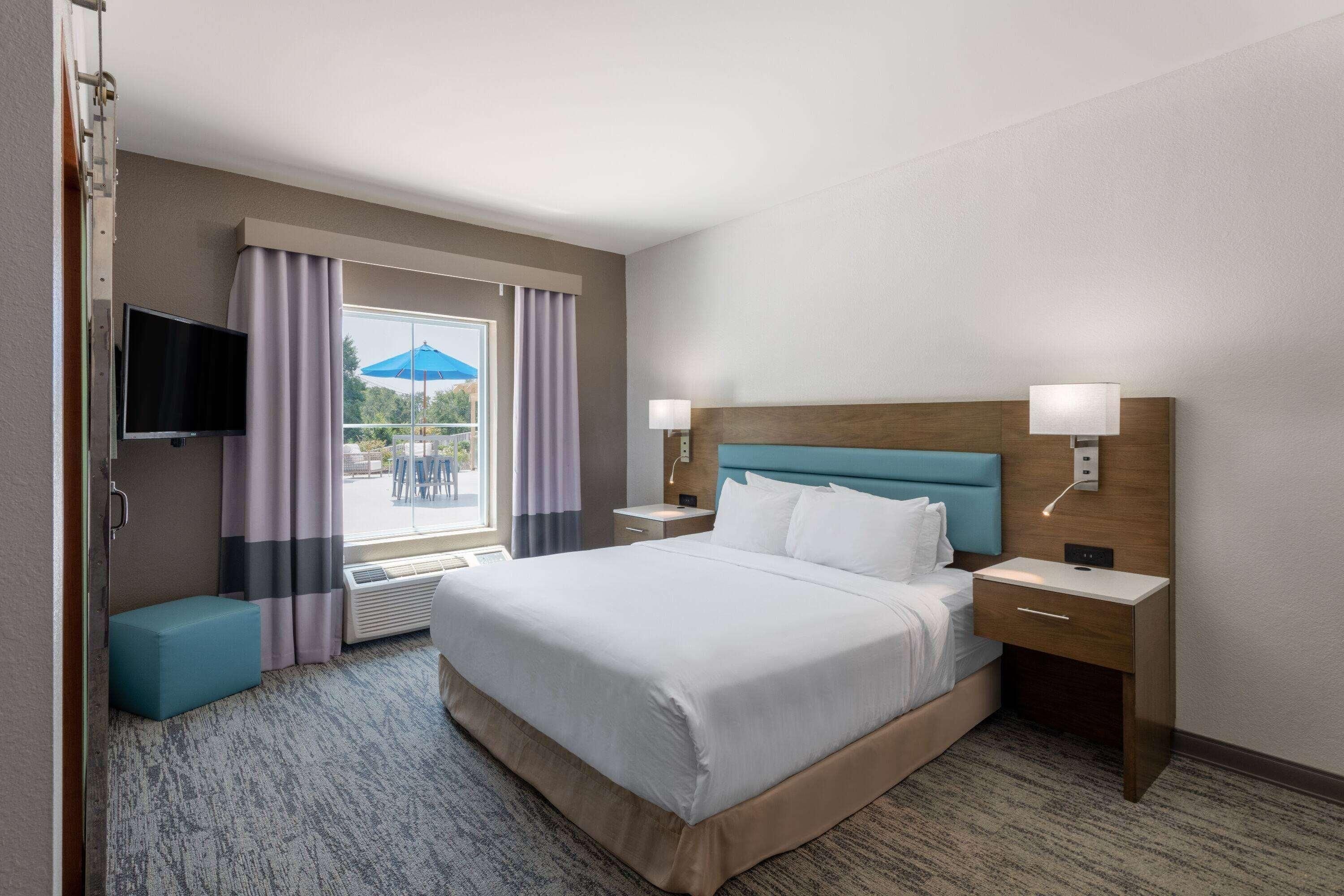 Quarto de hotel com uma cama grande, duas mesas de cabeceira, televisão e janela com cortinas.