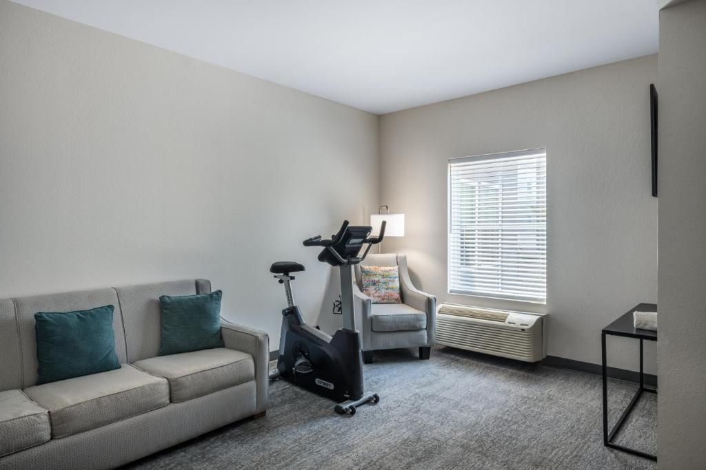 A room with a sofa, an exercise bike, a chair, a table, and a window with blinds.