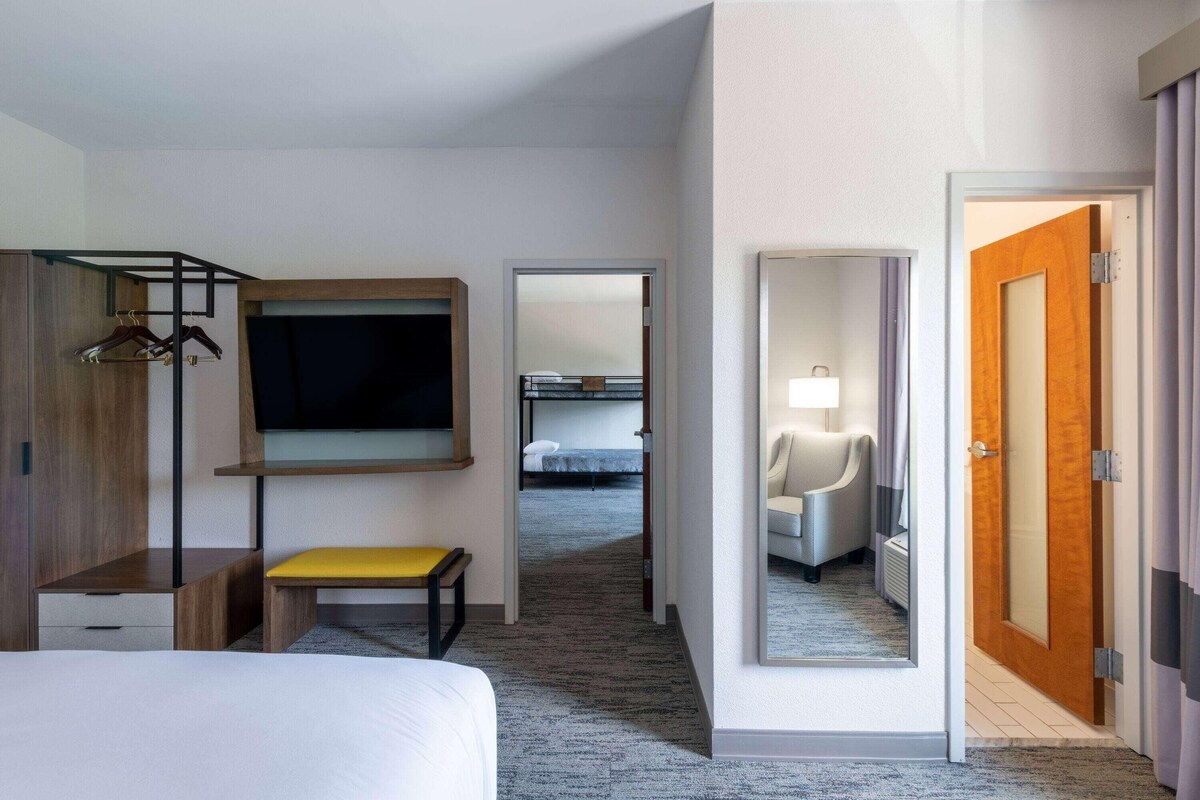 A hotel room with a bed, a bench, a TV, a mirror, a door, and a hallway leading to another room.