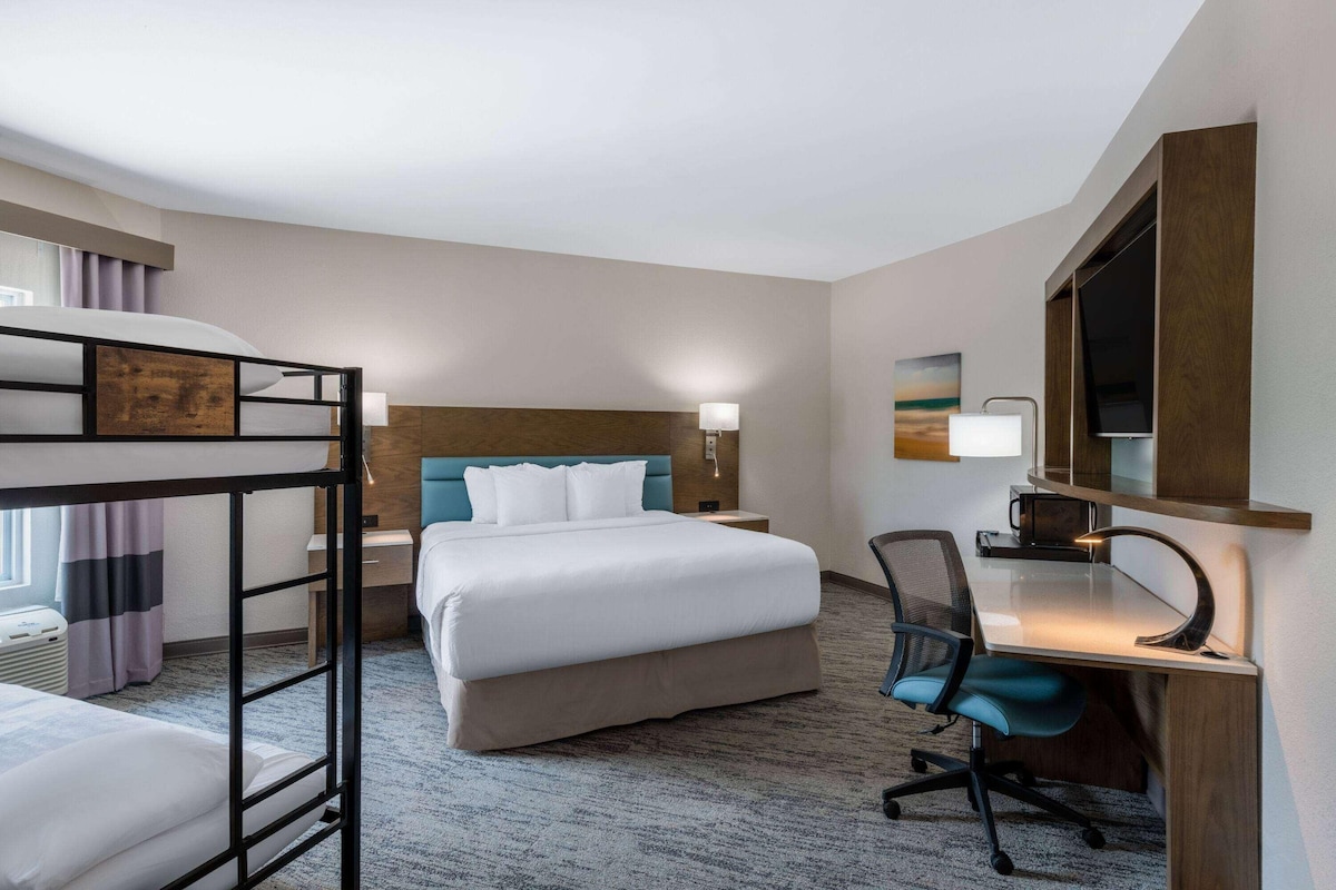 A hotel room with a bunk bed, a desk with a chair, a desk lamp, and a painting on the wall.