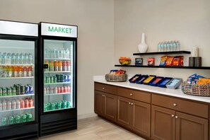 A kitchenette with a refrigerator labeled 'MARKET' stocked with beverages, a countertop with snacks, and a white vase on a shelf. - Wingate by Wyndham Williamsburg (Williamsburg)