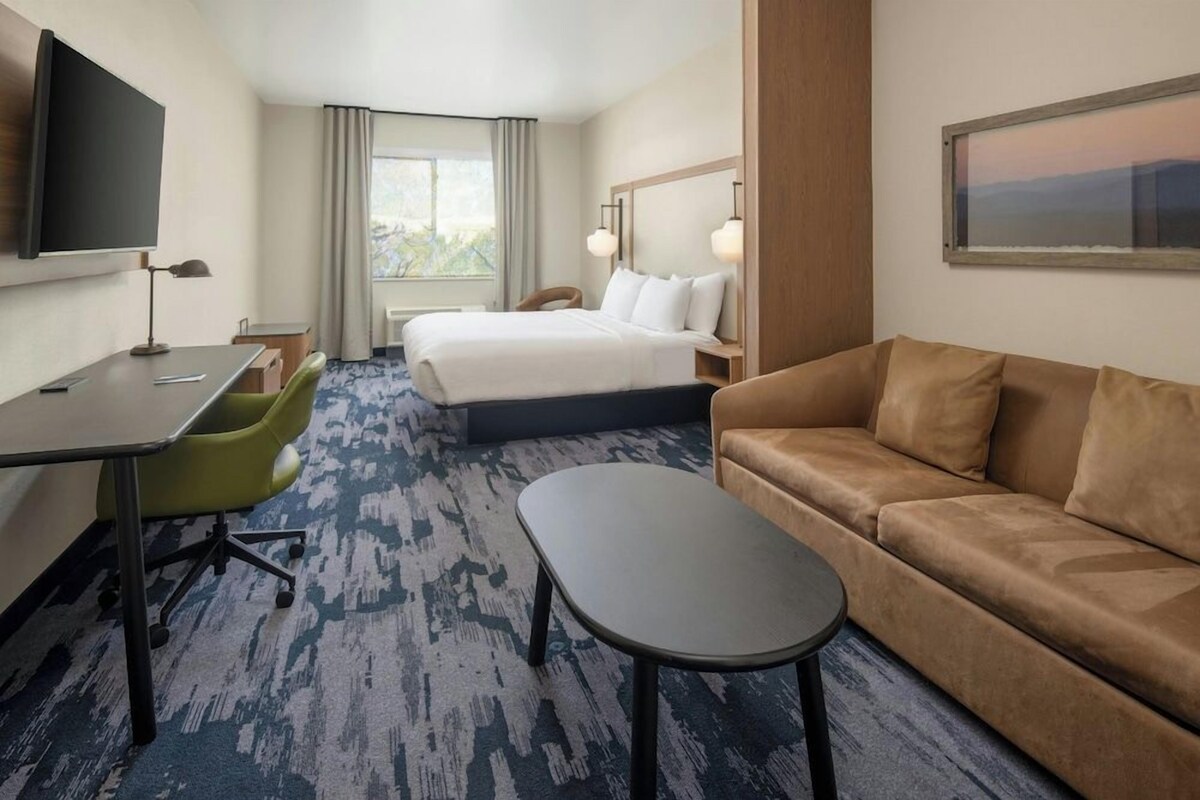 A hotel room with a bed, a sofa, a desk with a chair, a TV, and a window with curtains.