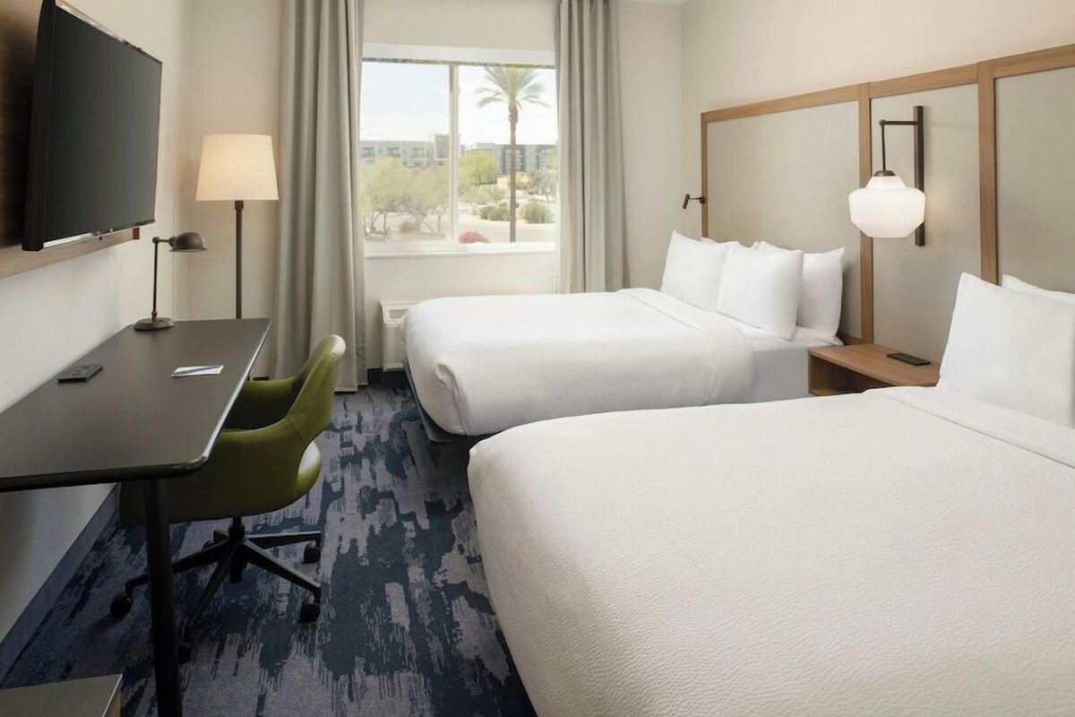 A hotel room with two beds, a desk, a chair, a TV, and a window with a view of palm trees.