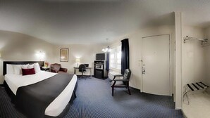 A hotel room with a large bed, a desk, a chair, a TV, a bathroom with a sink and mirror, and a shower area. - Evergreen Studios (Sheridan)