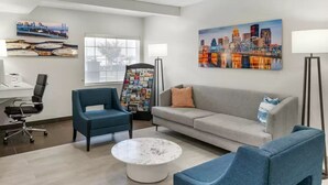 A modern waiting area with a blue armchair, a grey sofa, a coffee table, and a cityscape painting on the wall.