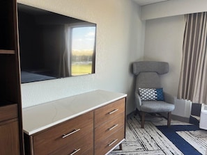 Habitación de hotel con televisor de pantalla plana montado en la pared, cómoda de madera con cajones, sillón gris con cojín estampado y alfombra decorativa en el suelo.