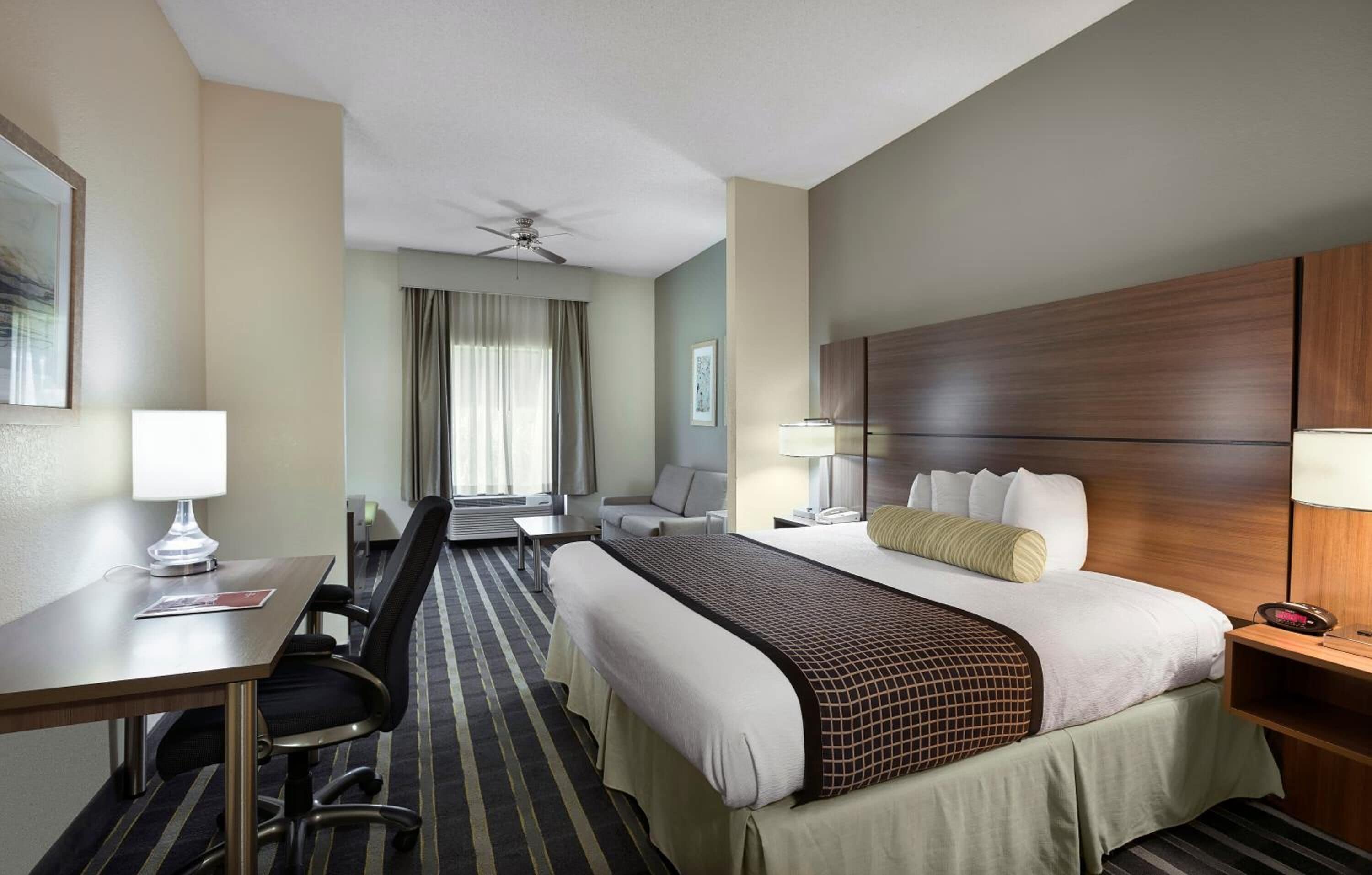 Une chambre d’hôtel comprenant un lit, un bureau, une chaise, une table de chevet, une lampe, un ventilateur de plafond et une fenêtre avec des rideaux.
