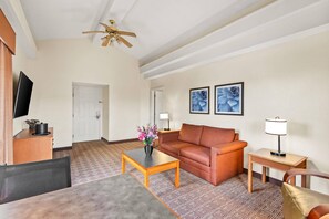 A hotel room with a sofa, a coffee table, a TV, a ceiling fan, and a patterned carpet. - Econo Lodge Augusta North - Washington Road (Augusta)