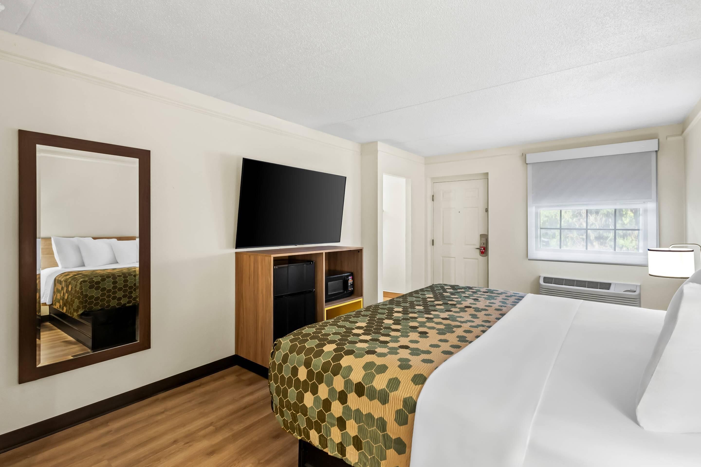 Une chambre d’hôtel équipée d’un lit, d’une télévision, d’un miroir, d’une fenêtre avec des stores et d’une porte.