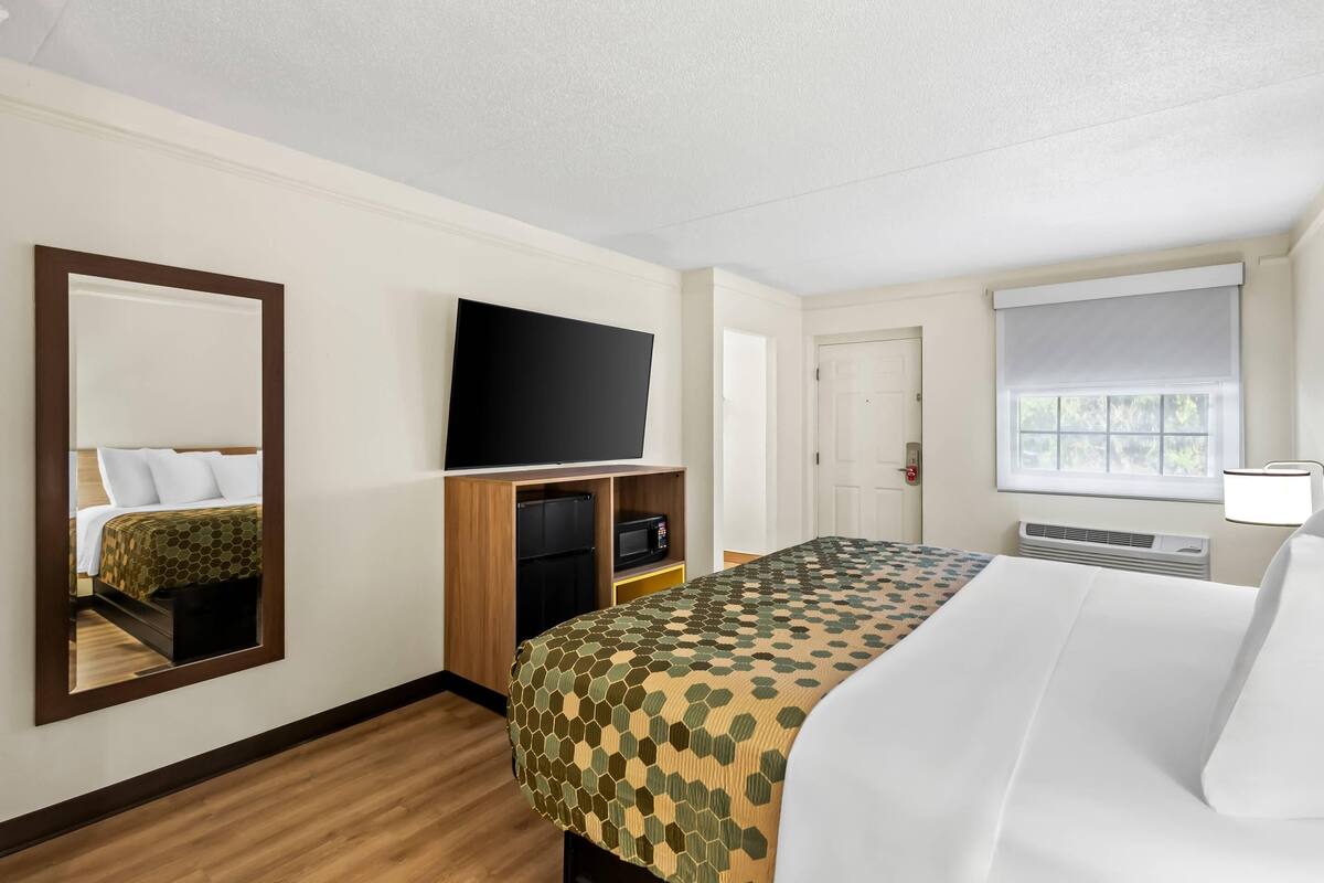 Habitación de hotel con cama, televisor, espejo, ventana con persianas y puerta.