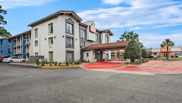 Un hotel de varios pisos con alfombra roja en la entrada, un área exterior techada y un cielo azul despejado.