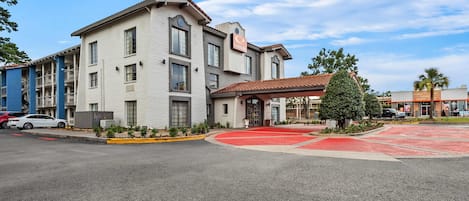 Un hôtel à plusieurs étages, avec un tapis rouge à l’entrée, un espace extérieur couvert et un ciel d’un bleu limpide.