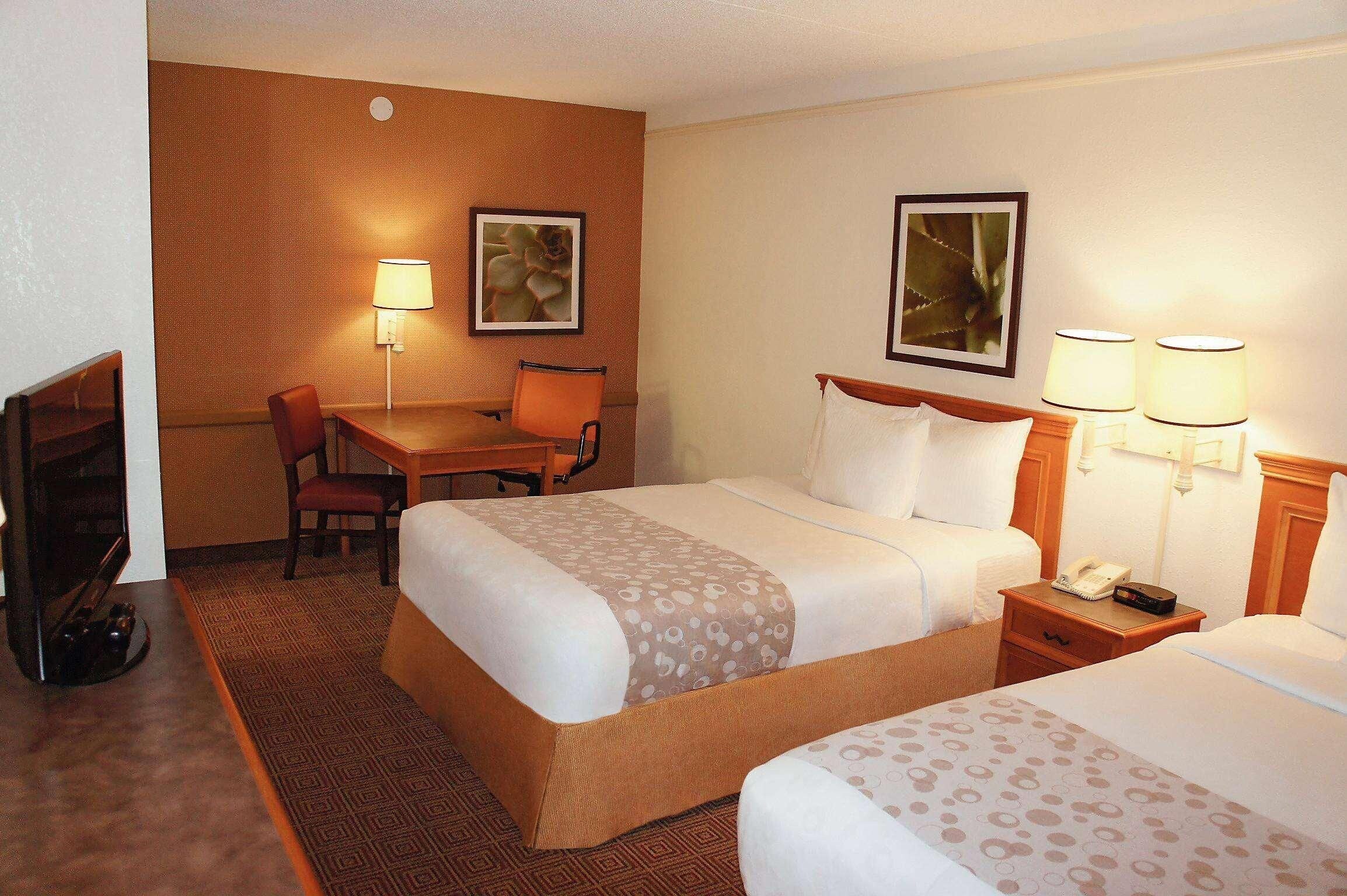 A hotel room with two beds, a desk, a chair, a TV, and two wall-mounted lamps.
