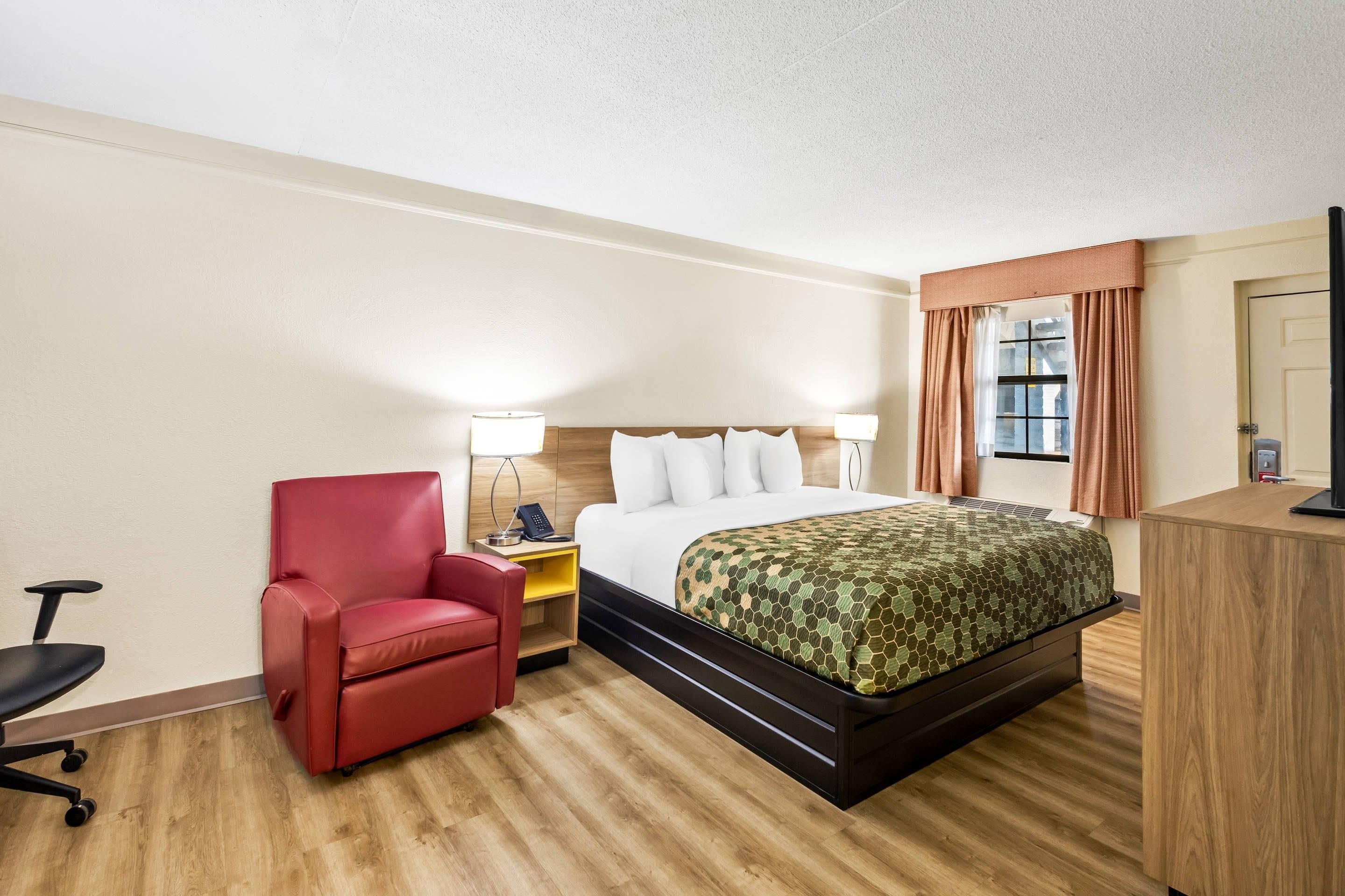 A hotel room with a bed, a red chair, a desk, a lamp, and a window with curtains.