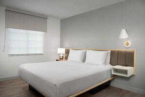 A neatly made bed in a hotel room with a headboard, a bedside table with a lamp, and a window with blinds. - Towneplace Suites by Marriott Birmingham South (Birmingham)