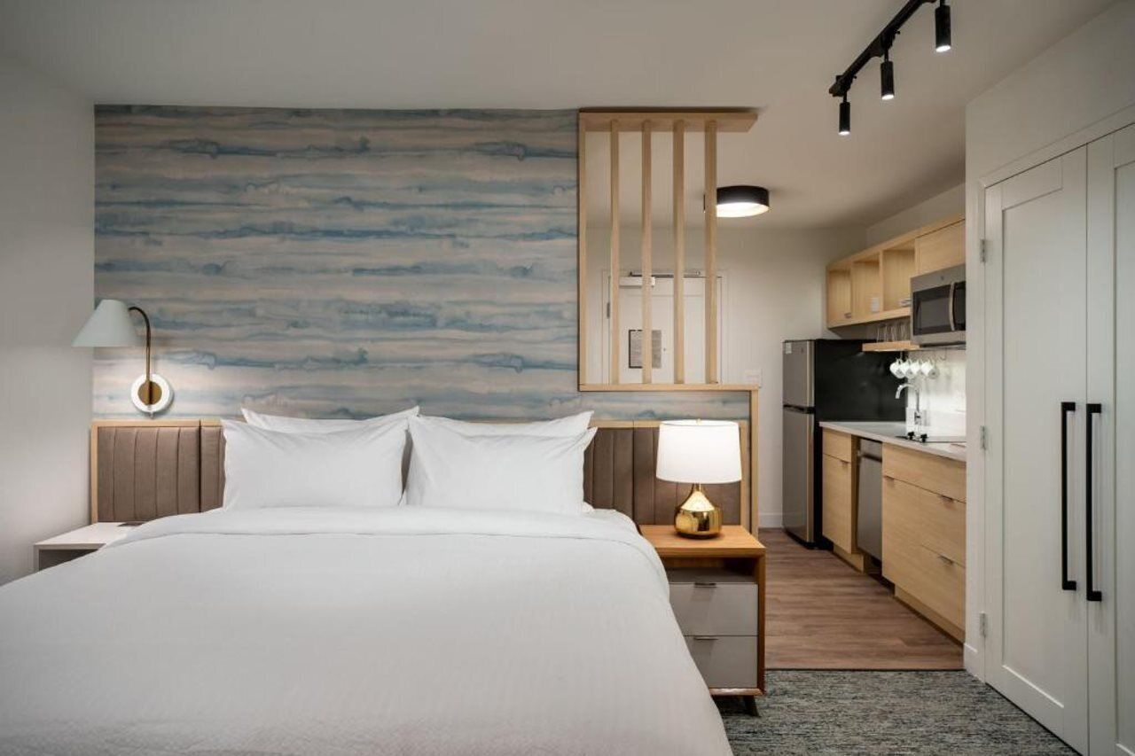 A hotel room with a bed, bedside lamp, and a view of the kitchen.