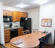 Cocina compacta con armarios de madera, un refrigerador negro, microondas y cafetera. Hay una mesa de comedor con sillas y un tablón de anuncios en la pared.