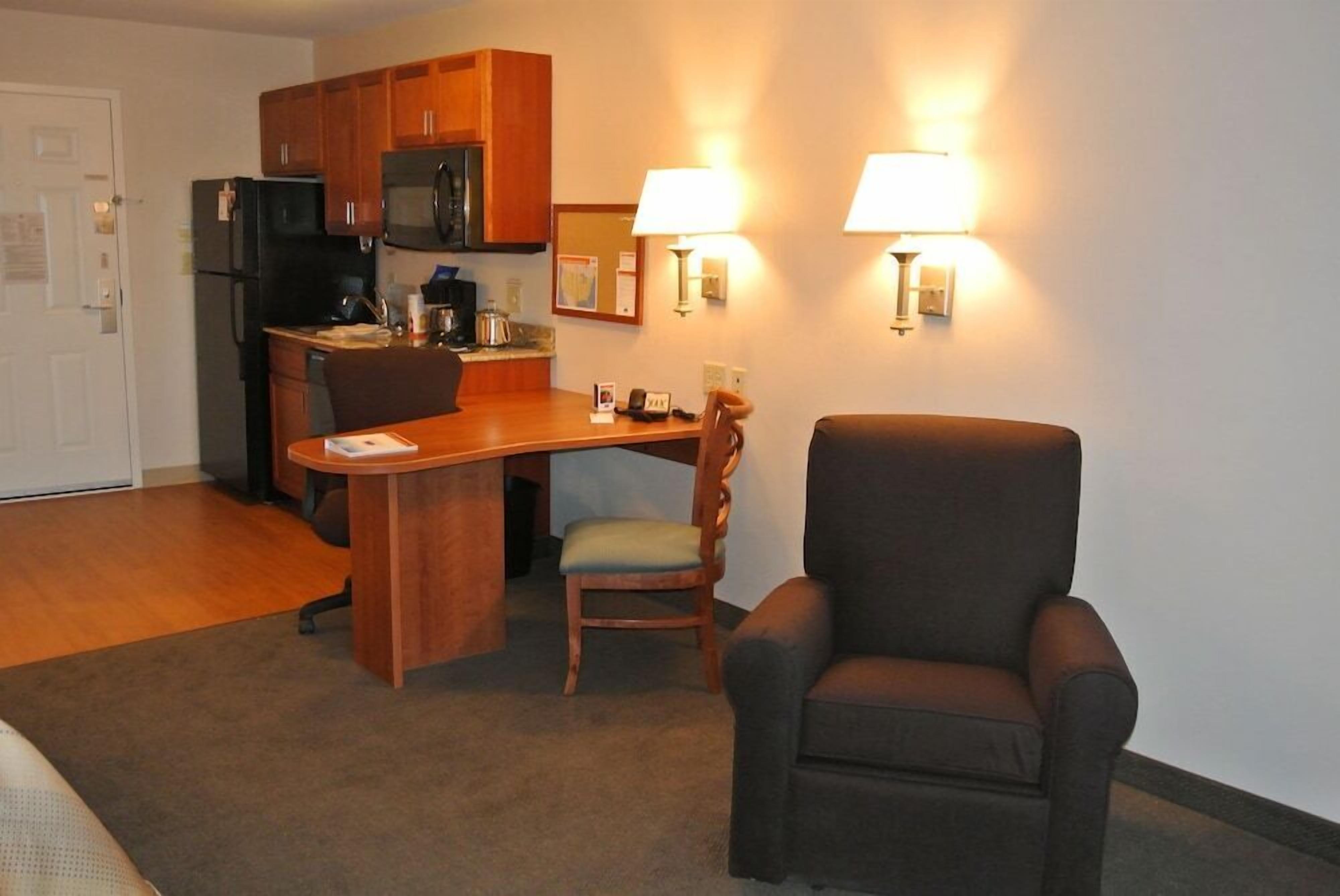 A compact kitchen with wooden cabinets, a microwave, and a coffee maker. There is a dining area with a wooden table and two chairs. Two wall-mounted lamps provide lighting.