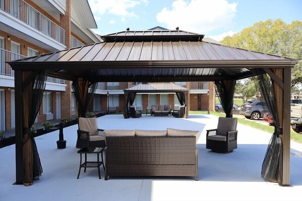Un área de estar al aire libre techada, con una pérgola, mobiliario de mimbre y vistas a un edificio al fondo.