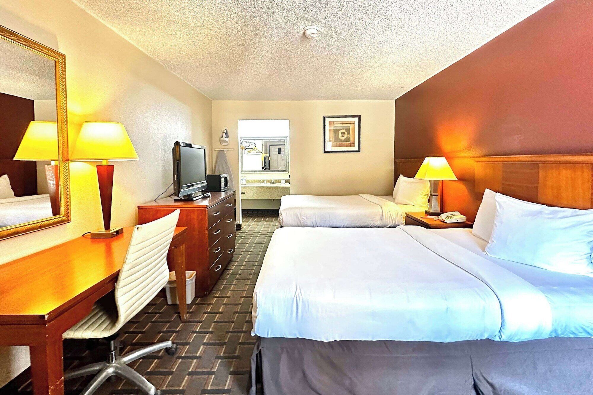 Habitación de hotel con dos camas, un escritorio con lámpara, cómoda, televisor y baño a la vista al fondo.