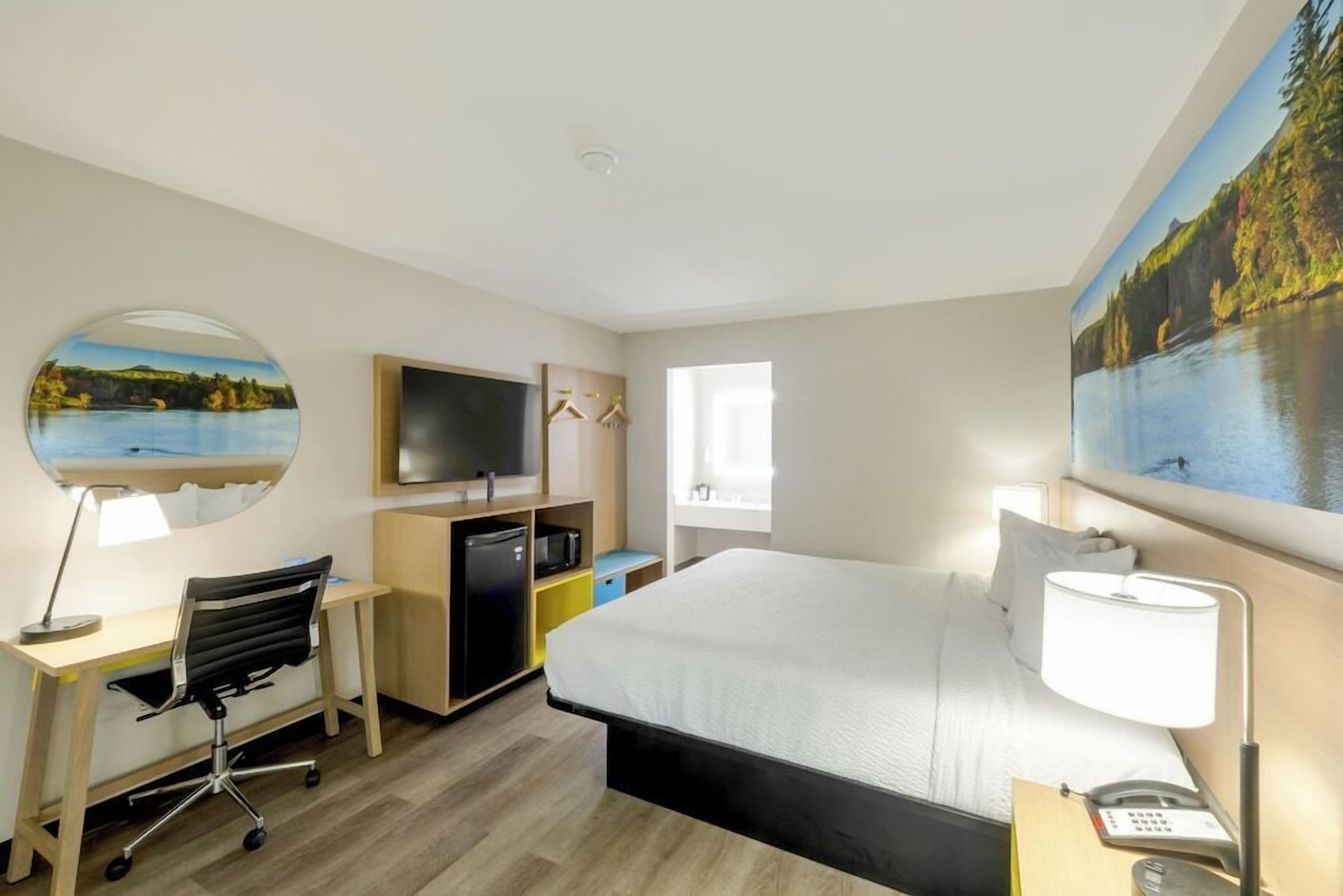 A hotel room with a bed, desk, chair, TV, and a painting of a lake.