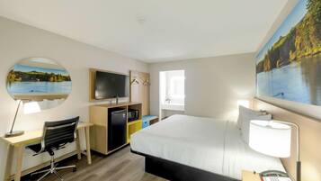 A hotel room with a bed, desk, chair, TV, and a painting of a lake.