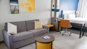 A hotel room with a grey sofa, a small round table, a desk with a chair, a bed, and a wall-mounted lamp. - Comfort Suites Baton Rouge South I-10 (Baton Rouge)
