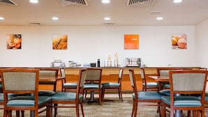 A hotel dining area with wooden tables and chairs, a counter with a coffee machine, and wall art. - Comfort Suites Baton Rouge South I-10 (Baton Rouge)