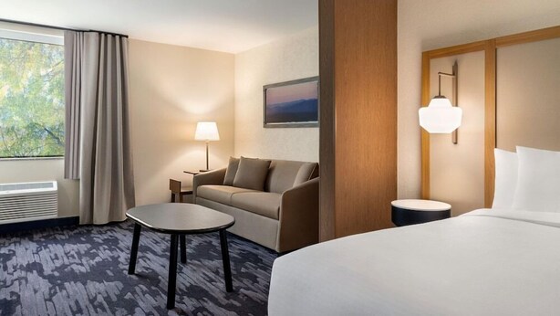 A hotel room with a bed, a sofa, a small table, a lamp, a picture on the wall, and a window with curtains. - Hotel Monona (Madison)