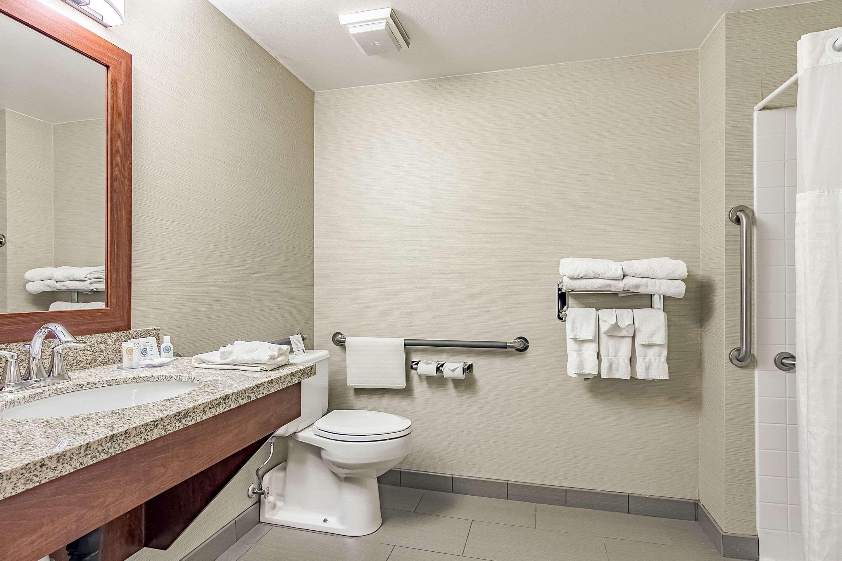 A modern bathroom with a granite countertop sink, a white toilet, a towel rack with towels, a handrail, and a shower area.