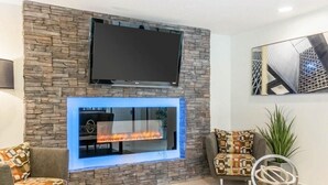 A living room with a stone wall, a flat-screen TV, a fireplace, two patterned armchairs, and a coffee table. - Fairbridge Inn Express Indianapolis Northwest (Indianapolis)