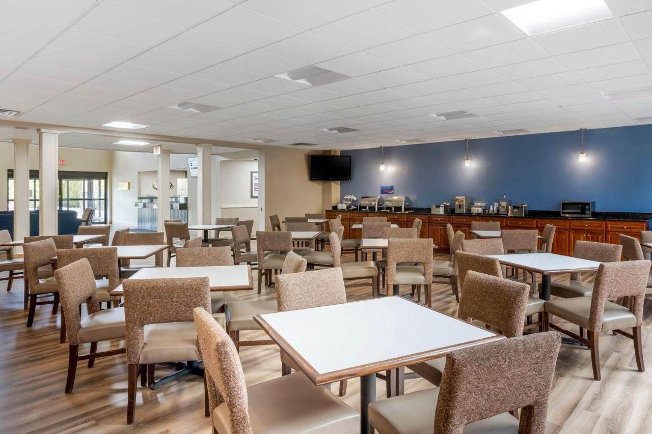 A hotel dining area with tables and chairs, a kitchen in the background, and a television on the wall.