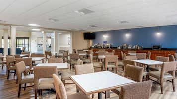 Une salle à manger d’hôtel avec des tables et des chaises, une cuisine à l’arrière-plan et un téléviseur accroché au mur.