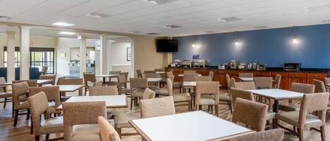 A hotel dining area with tables and chairs, a kitchen in the background, and a television on the wall.