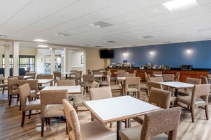 Comedor de hotel con mesas y sillas, una cocina al fondo y un televisor en la pared.