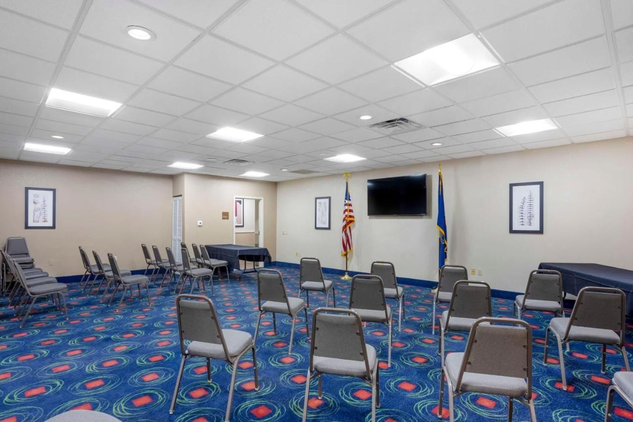 Un salón de conferencias con alfombra azul, filas de sillas, una mesa, banderas y una pantalla de televisión.