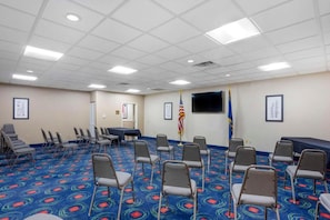 Een congrescentrum met een blauw tapijt, rijen stoelen, een tafel, vlaggen en een televisiescherm.