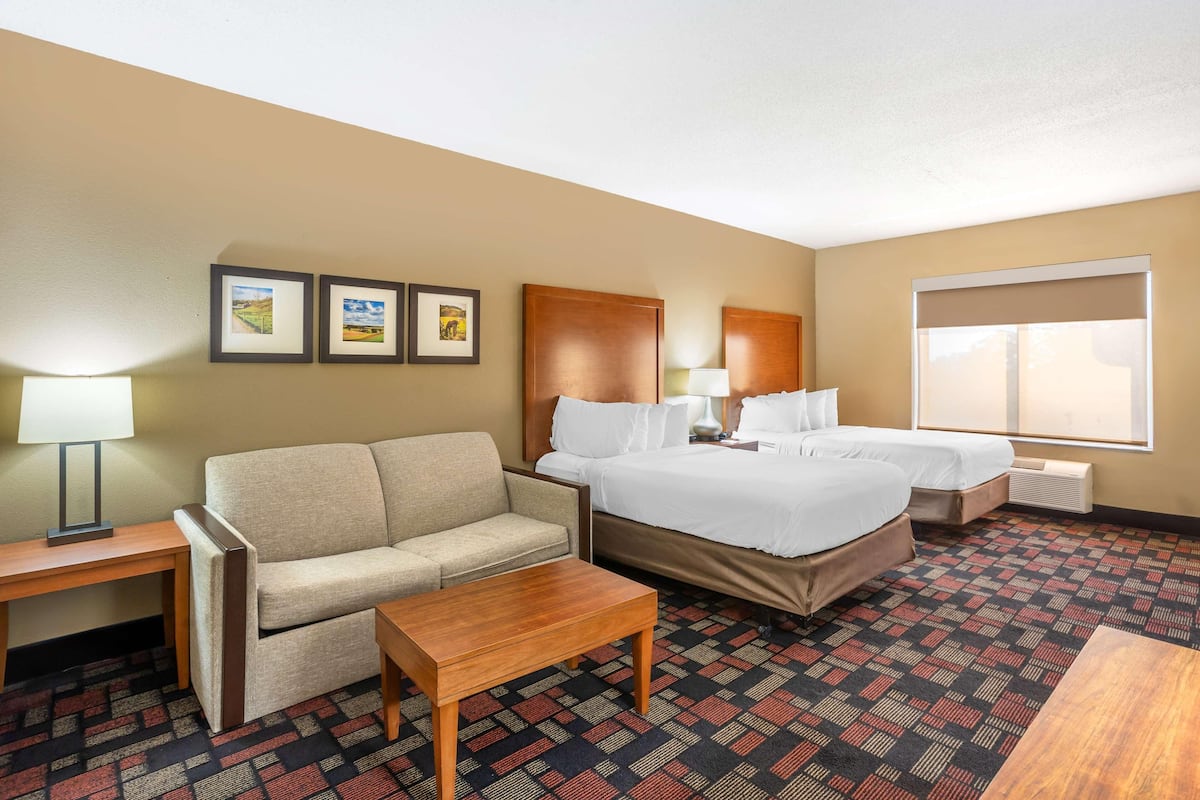 A hotel room with a sofa, two single beds, a wooden table, a lamp, and framed pictures on the wall.