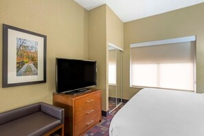 Ein Hotelzimmer mit einem Bett, einem Fernseher auf einer Holzkommode, einem gerahmten Bild an der Wand und einem Fenster mit Jalousien.