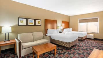 A hotel room with a sofa, two single beds, a wooden table, a lamp, and framed pictures on the wall.