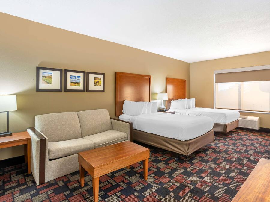 A hotel room with a sofa, two single beds, a wooden table, a lamp, and framed pictures on the wall.