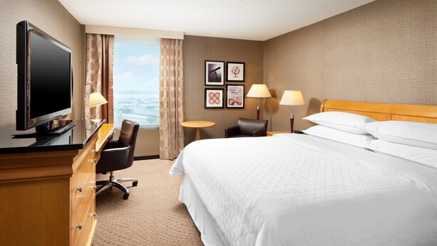 A hotel room with a large bed, a desk with a TV, two chairs, a small table, and a view of the city. - Cleveland Airport Hotel (Cleveland)