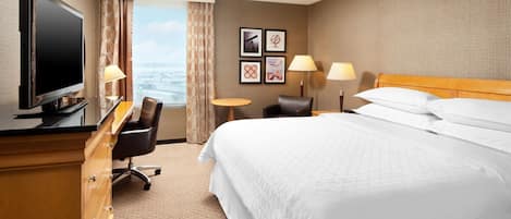 Ein Hotelzimmer mit einem großen Bett, einem Schreibtisch mit Fernseher, zwei Stühlen, einem kleinen Tisch und Blick auf die Stadt.