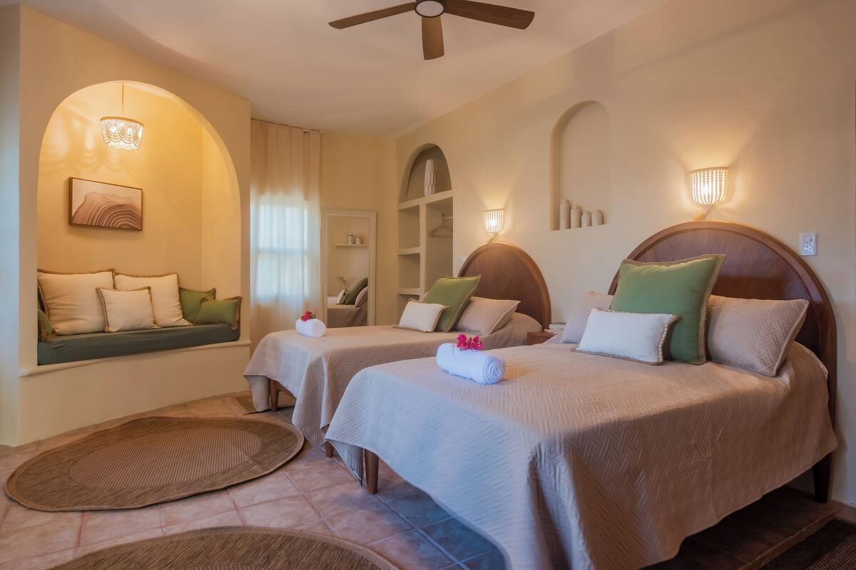 A hotel room with two beds, a sitting area with a bench, a ceiling fan, and wall-mounted lights.