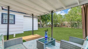 En overdækket terrasse med fletmøbler, et glasbord og udsigt til en velholdt græsplæne og træer.