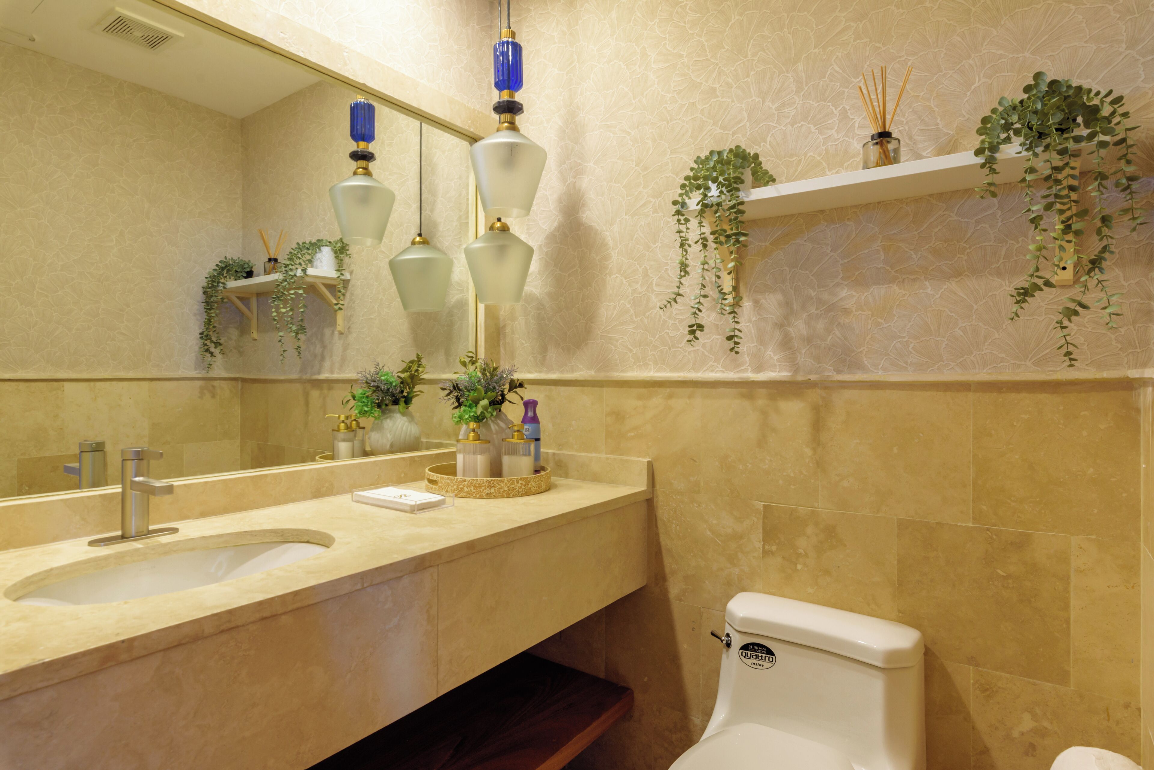 Ein Badezimmer mit beiger Fliesenwand, einem Waschbecken inklusive Spiegel, einer Toilette sowie Regalen mit Pflanzen und Pflegeprodukten.