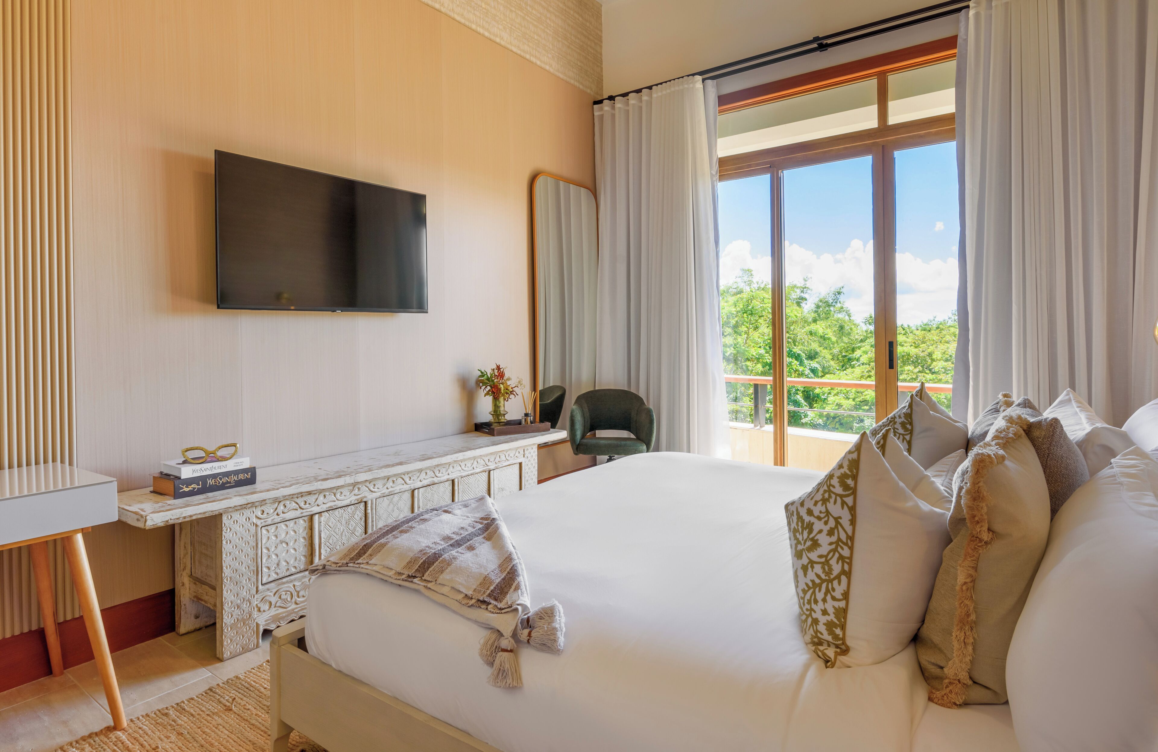 A hotel room with a large bed, a TV mounted on the wall, a desk with a chair, and a view of greenery through the window.