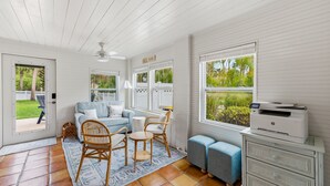 Une pièce avec un plafond blanc, un ventilateur de plafond, un canapé, une chaise, une petite table, une imprimante et une vue sur la verdure à l’extérieur.