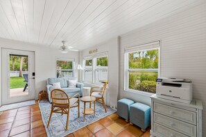 A room with a white ceiling, a ceiling fan, a sofa, a chair, a small table, a printer, and a view of greenery outside. - Sunny Palm Retreat • Fenced Yard • Outdoor Dining (Jensen Beach)